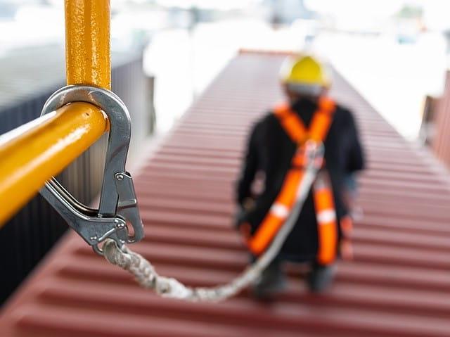 Metal harness clip attached to yellow guardrail is attached by a rope to man in construction hat wearing orange safety harness